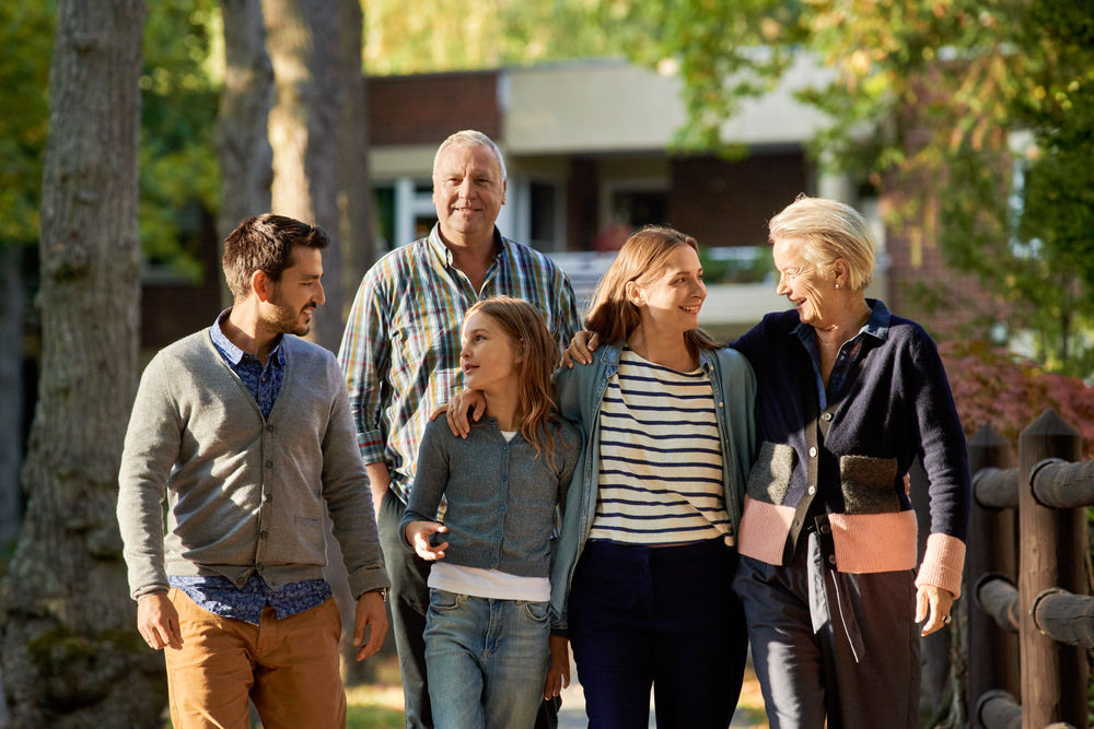 Eine Familie geht spazieren: Oma, Opa, Vater, Mutter, Tochter, im Hintergrund Bäume und ein Mehrfamilienhaus.